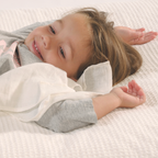 A toddler wearing a light grey long-sleeve shirt holding a white cuddle blanket lying on a textured white waffle-weave mattress.