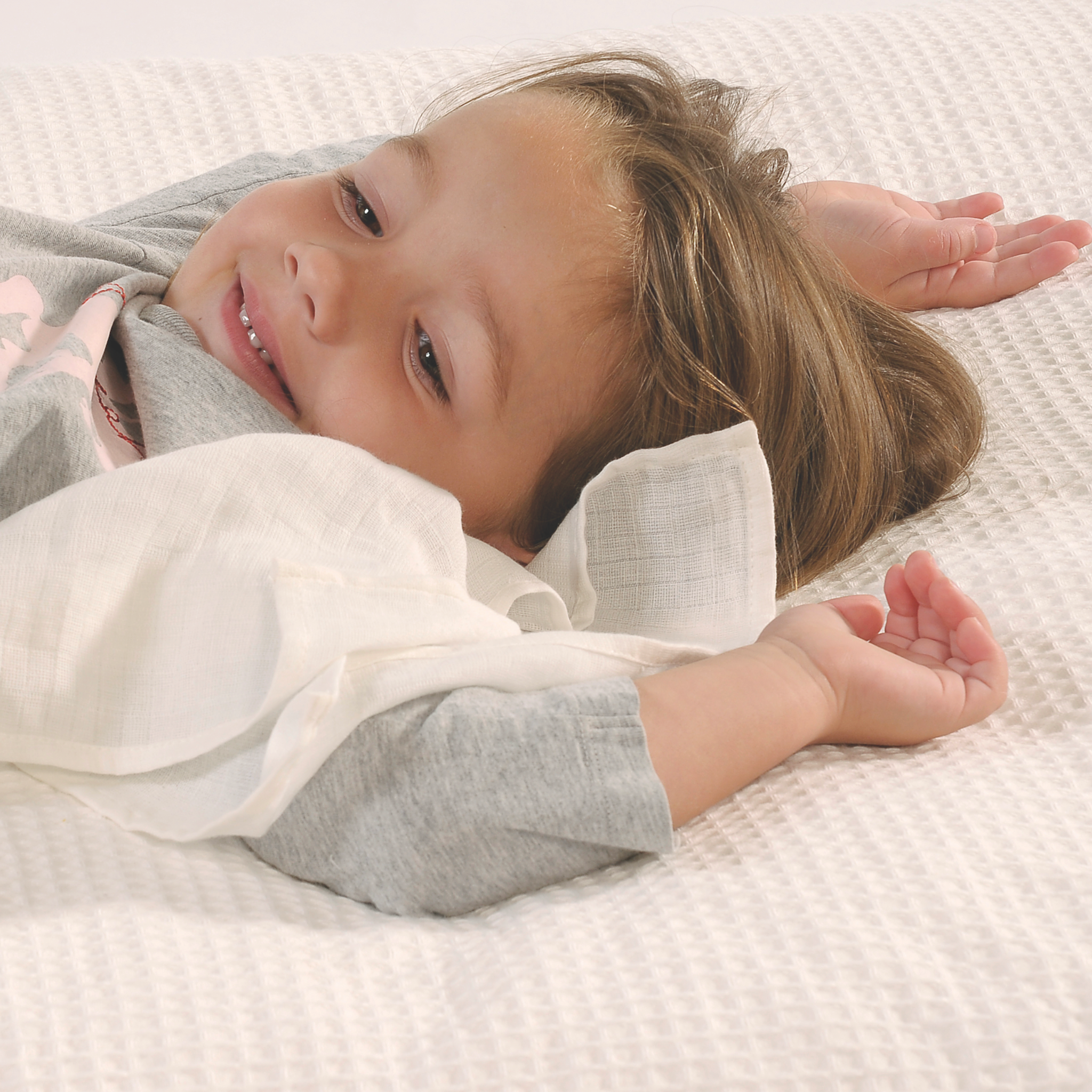 A toddler wearing a light grey long-sleeve shirt holding a white cuddle blanket lying on a textured white waffle-weave mattress.