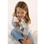 A young child with light brown hair sits on a white textured bed, holding a white bear my luvvie baby comforter. The child is wearing a light grey long-sleeve shirt and blue jeans.
