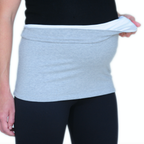A close-up of a person wearing a light grey maternity tummy band over a black top and black leggings, holding the fabric out to show its stretch.